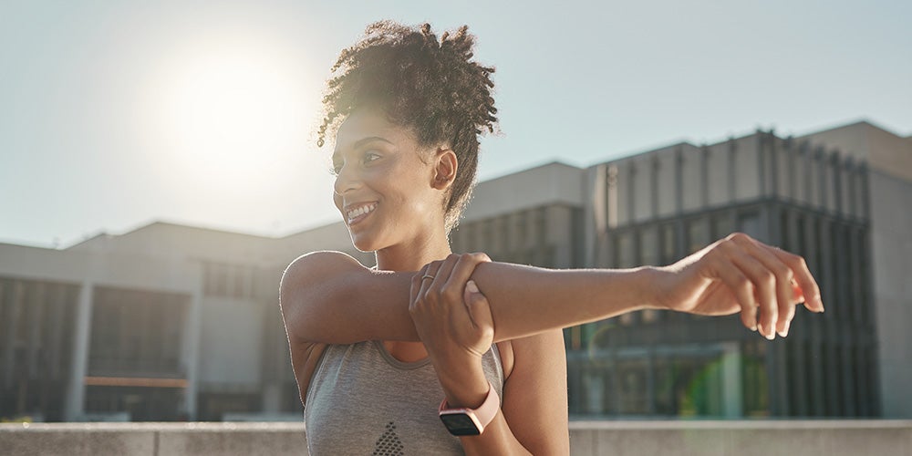 Smiling woman with curly hair stretching her arm outdoors on a sunny day, wearing an athletic top and smartwatch.