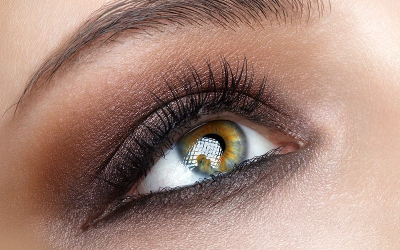 Close-up of a hazel eye with brown eyeshadow and long dark lashes, reflecting a grid pattern in the pupil.