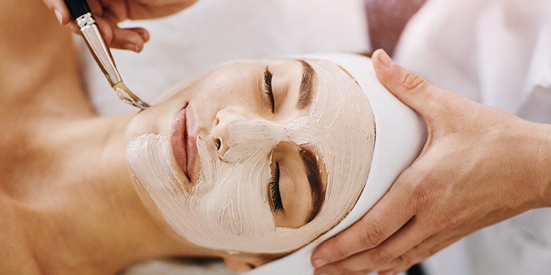 Woman with closed eyes receiving a facial, a white mask being applied to her face with a brush by an unseen person.