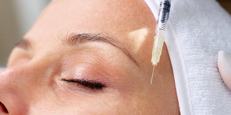 Close-up of a woman's face receiving an injection near her temple, with a syringe needle positioned near her skin.