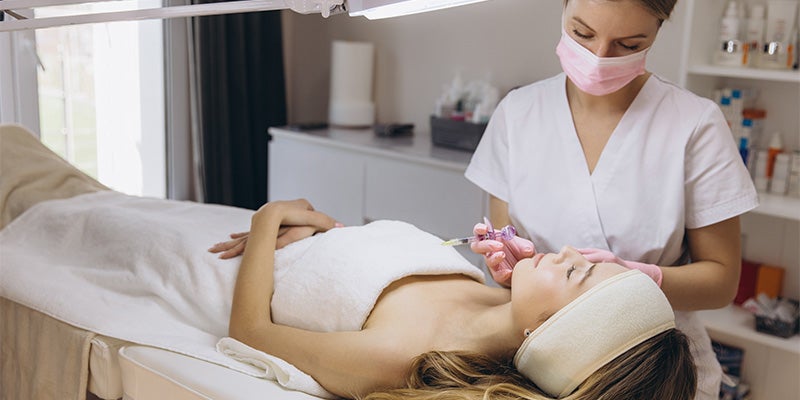 Esthetician in a mask and gloves injecting a woman's face on a spa bed.