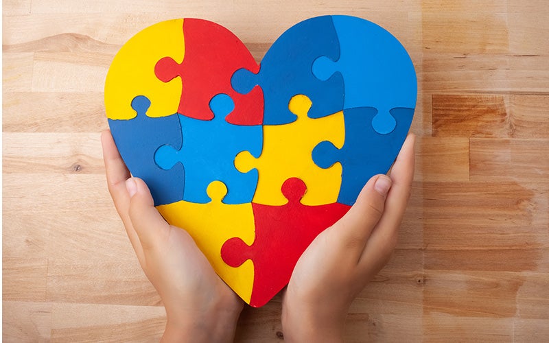 Hands hold a heart-shaped puzzle made of red, blue, and yellow pieces, symbolizing autism awareness.