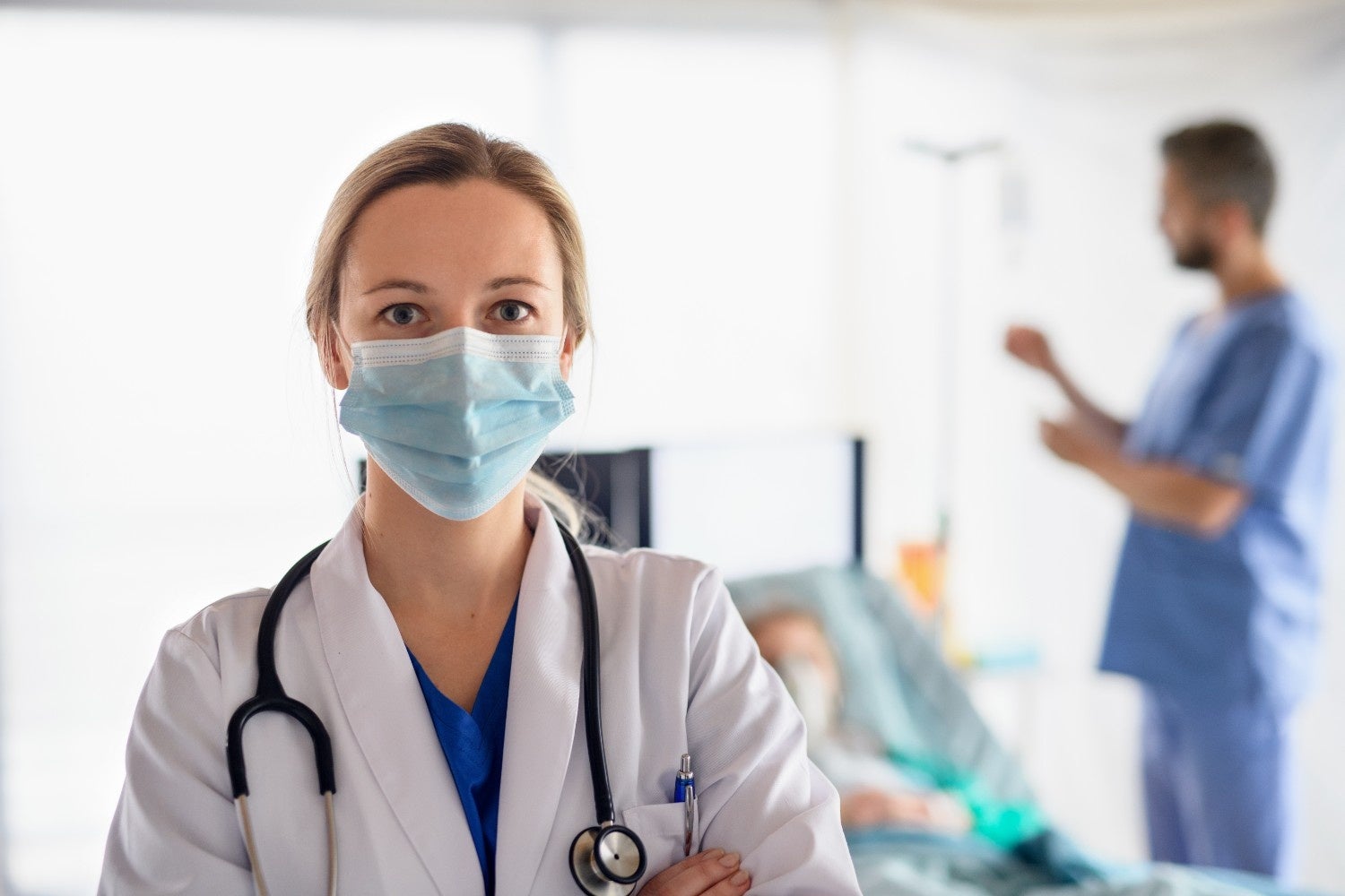 Female doctor in a mask and white coat with a stethoscope, looking at the camera in a hospital room.