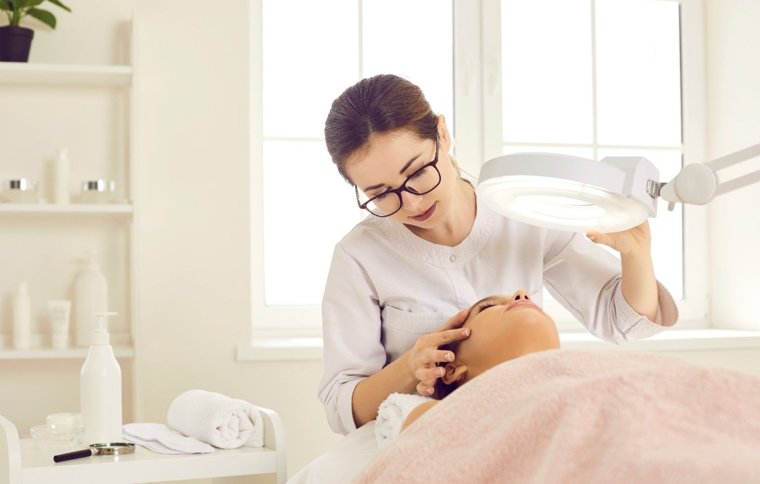 Esthetician examines client's face under a magnifying lamp in a bright beauty salon.