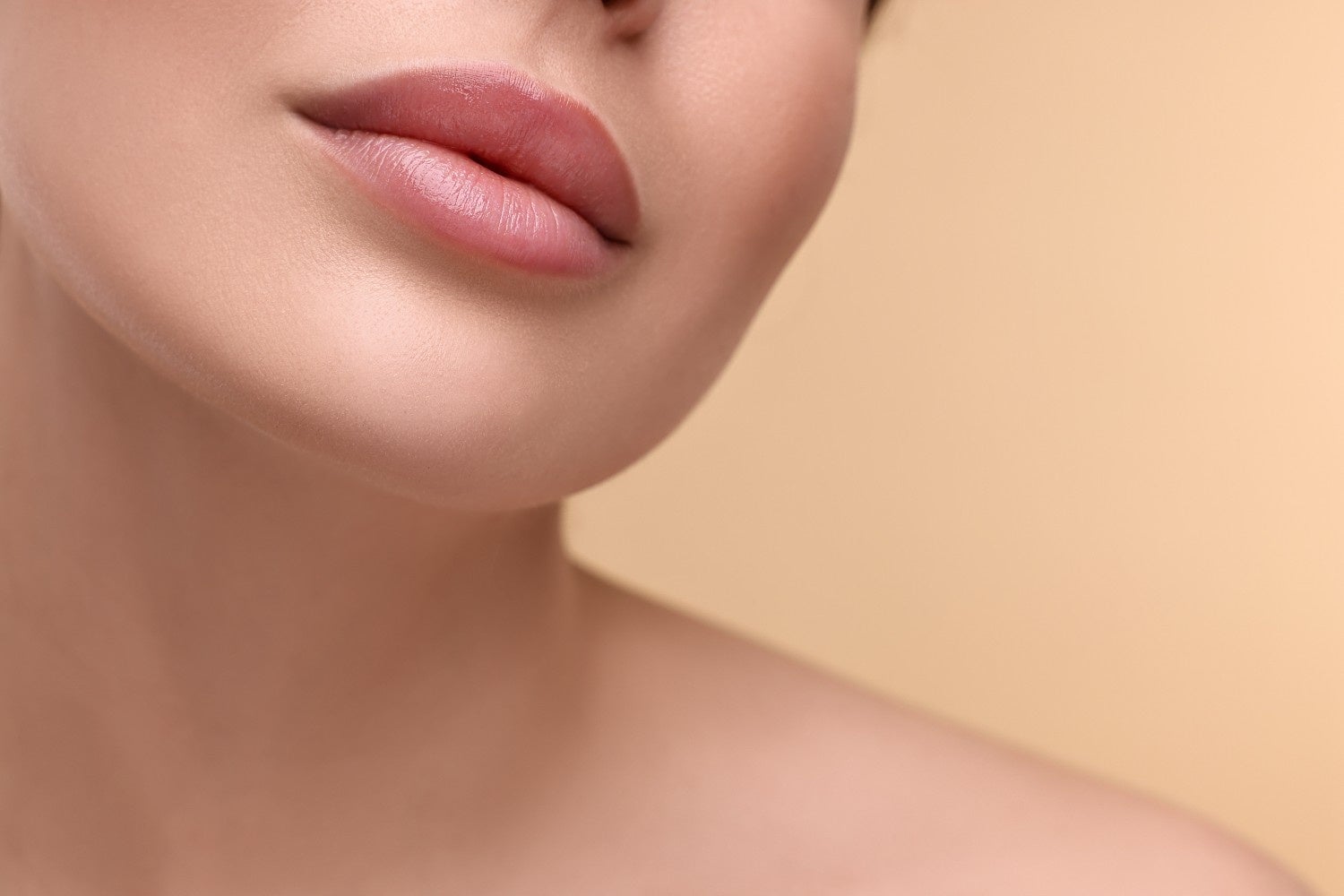 Close-up of a person's lower face and shoulder, showing plump, pink lips and smooth skin against a beige background.